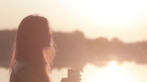 close-up portrait attractive young girl looks at the sunrise on the river and drinking coffee from a thermocup
