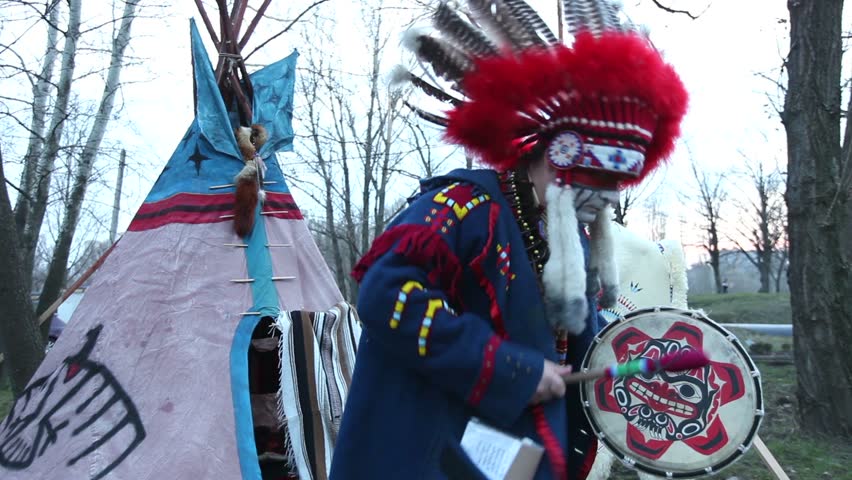 North American Indian in full dress, A man in a suit Indian dancing with a tambourine on the street in front of wigwam, day,