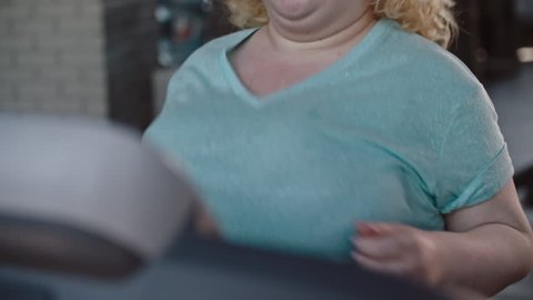 Large woman running on a treadmill and then walking, wiping her face with towel and drinking water