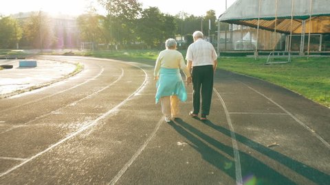 Senior couple taking a walk along the running track in summer. Healthy retirees enjoying morning walking on the stadium with camera lens flare.