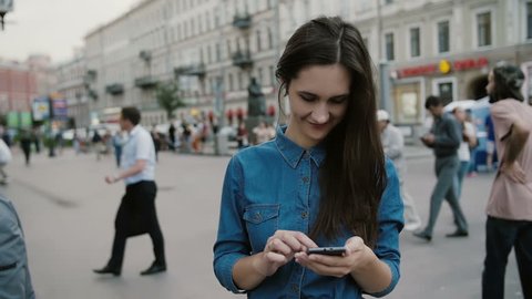 Modern technology. Beautiful brunette young woman in a denim dress using her smartphone in a busy street. Slow mo
