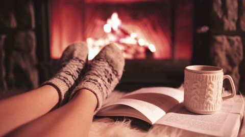 Woman Reading a Book by the Cozy Fireplace. 4K Stabilized shot. Young Unrecognizable Girl enjoying a Storybook by the warm Fireside With a Cup of Hot Drink in Ornamented Cup. Relaxed holiday evening.