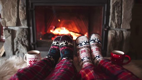 Feet in woollen socks by the Burning Christmas Cozy Fireplace. 4K. Couple relaxes by warm fire with cup of hot drink and warming up their feet. Winter and Christmas holidays concept.