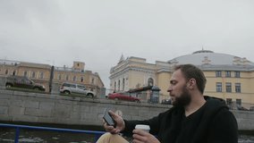 Man on a boat tour drinking coffee, sightseeing in St Petersburg. Holding smartphone in hand. City architecture, slow mo
