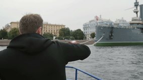 Sightseeing in St Petersburg. Man uses smartphone, takes photos of the Cruiser Aurora, passing by it on a boat, slow mo