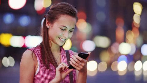 Woman using smart phone at night in city. 4K. Attractive young Woman texting, talking on smartphone outdoors. Tourist Millennial Girl with cellphone, blurred Night Busy Street lights background.