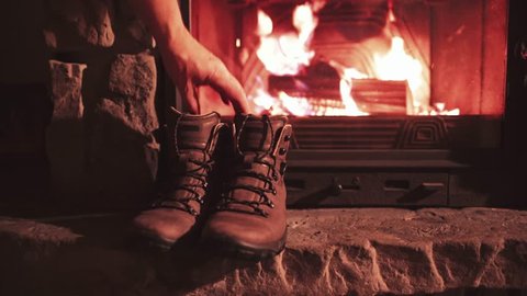 Man Hand takes Warm Winter Boots from a Shelf near Fireplace. 4K. Vintage folk boots near the Cozy Fireside. Shoes getting warm near the burning fire at home, in chalet. Autumn and Winter Concept