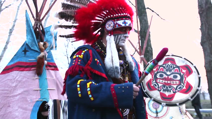 North American Indian in full dress, A man in a suit Indian dancing with a tambourine on the street in front of wigwam, day
