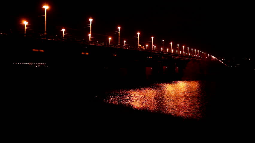 The night bridge in a city with passing cars and light reflections on river water