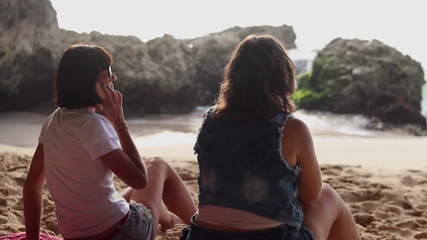 Pretty women with glasses sitting on the beach near the ocean, talking, laughing and smiling. One woman using smartphone. Girls vacation on Bali island, Indonesia. The best friends. Back view.