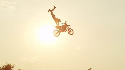 SLOW MOTION: Extreme pro motocross rider riding fmx motorbike, jumping huge jump performing dangerous stunt against the sky. Professional motocross biker jumps big air trick over golden sunset sun