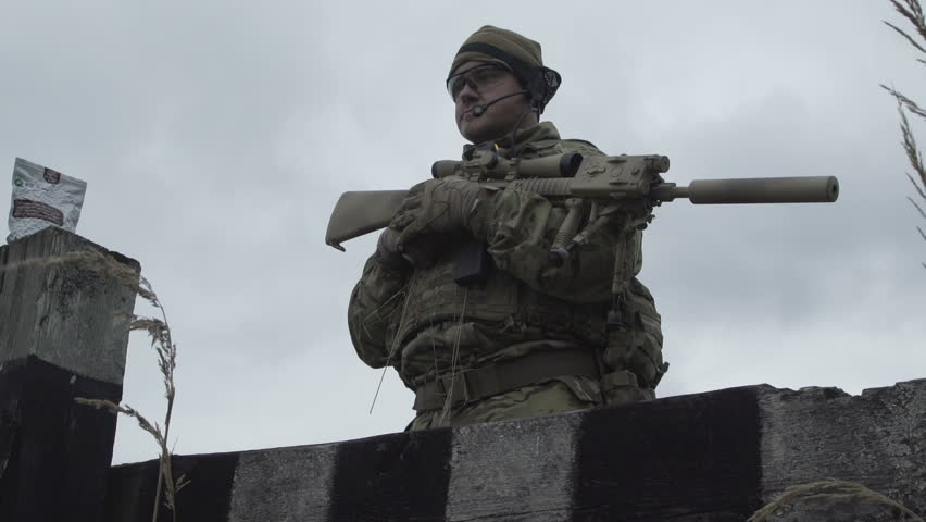 Military in an army uniform and hat standing in a field on a background of sky and grass. A soldier with a gun near the shelter