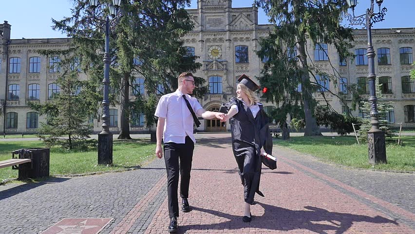 Happy Graduands Couple Walking by Cobblestone Alley and Holding Hands, Young Woman in Student Hat and Mantle. Slow Motion. Girl is Holding Rolled up Diploma Tied up With Red Tape. Couple of Students.