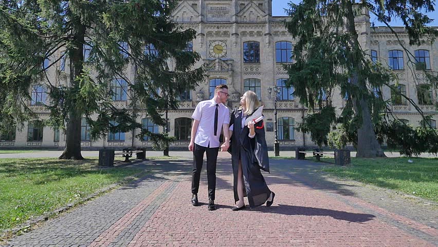 Happy Graduands Having Fun, Young Woman in Student Hat and Mantle Throws Her Hat Up, Catches it and Puts on Again. Slow Motion. Girl is Holding Rolled up Diploma Tied up With Red Tape. Couple of