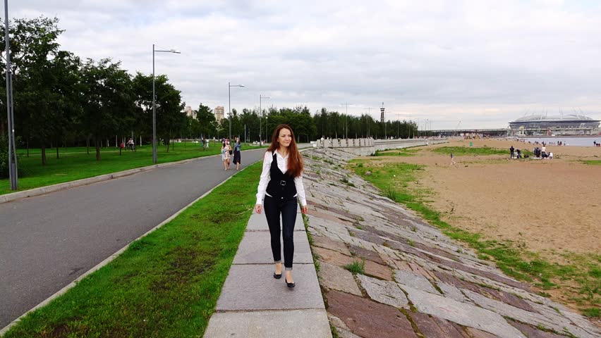 Thoughtful brunette woman go on shore front promenade in 300th Anniversary Park in Saint Petersburg. Pretty girl confidently walk against Gulf of Finland beach. People have rest on sand near water