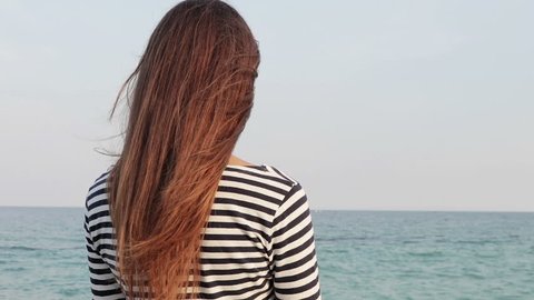 Young woman with long hair in a striped shirt looking at the sea, view from the back, slow motion