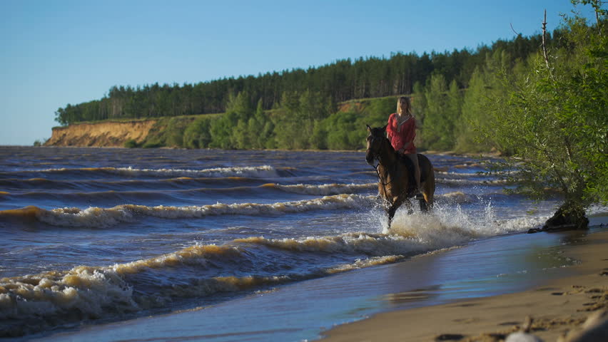 A beautiful blonde young woman riding a horse at a lake