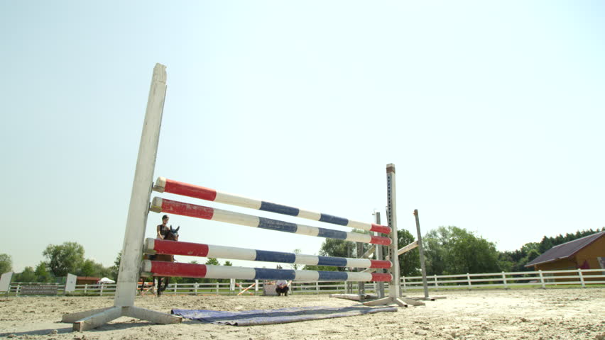 SLOW MOTION, CLOSE UP, LOW ANGLE: Horsegirl riding strong brown horse jumping the fence in sunny outdoors sandy parkour dressage arena. Competitive rider training jumping over obstacles in manege Royalty-Free Stock Footage #20075314