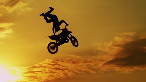 SLOW MOTION SILHOUETTE: Motocross rider riding fmx motorbike, jumping big air kicker performing extreme stunt. Professional biker jumps no footer knack knack trick over golden sunset sky above trees