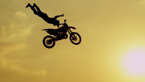 SLOW MOTION SILHOUETTE: Pro motocross rider riding fmx motorbike, jumping big air kicker performing extreme stunt. Professional biker jumps no hander trick over golden sunset sky above trees