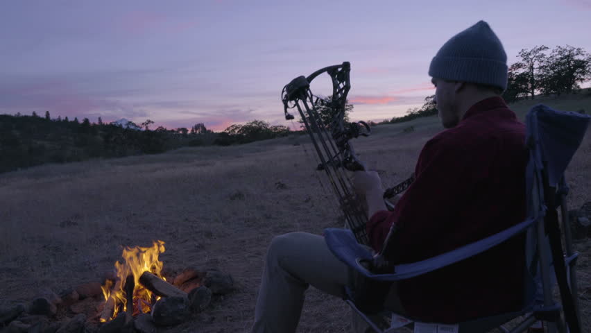 Man Sits In Camp Chair By Fire, At Sunset, And Checks Out His Hunting Bow