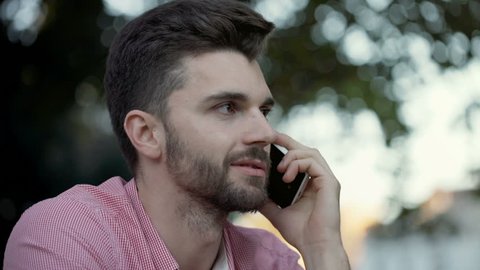 Calm man sitting outdoors and looking bored while talking on cellphone
