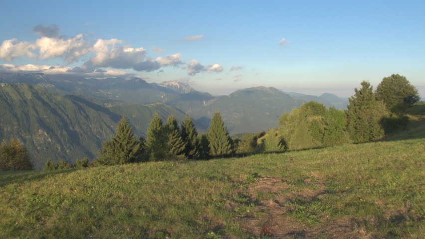 AERIAL: Flying above narrow road leading uphill to the rocky mountain ridge. Slope foothills, amazing vast lush forest and beautiful river flowing through stunning green valley and small town