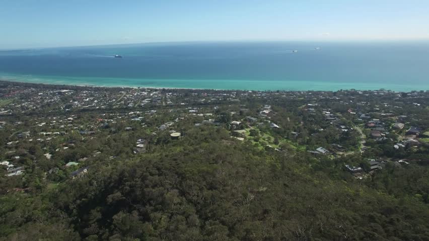 Forward flight from Seawinds Lookout high up on a hill towards Port Phillip bay with large cargo ship sailing across on hot summer day. Melbourne, Victoria, Australia