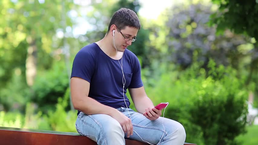 Young man sits on the bench in city park and speaks on smartphone. Man sits on the bench in public garden and uses mobile phone. Man communicates via smartphone Royalty-Free Stock Footage #20107291