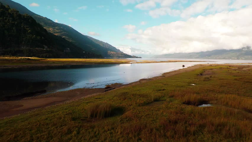 Aerial backwards tracking over clear alpine lake water reflecting the sky and mountains in Patagonia Chile with grasses and marsh in the foreground. 2K wide angle tracking landscape panorama shot.