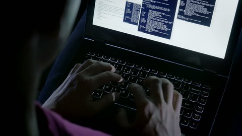 Close-up of hacker's hands typing codes on the keyboard. Young sly man trying to breach the system while giving bad commands in the dark.