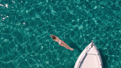 Aerial - Top view of a beautiful young woman jumping from a motor boat bow into crystal clear transparent water of a luxury travel destination