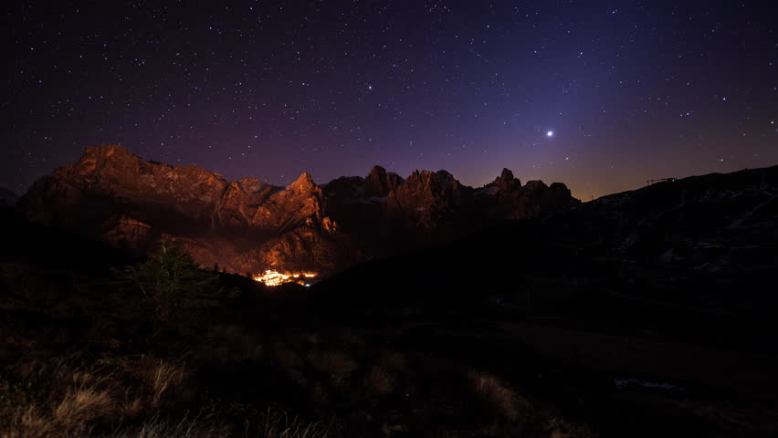 Sweeping dawn time-lapse of valley with a small village nestled at the base of the snowy mountain. The star-filled night sky turns golden as the sun rises through frame. Several shooting stars.