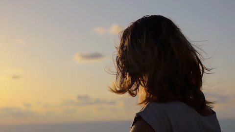lonely young woman looking out to sea at sunset as the wind moves the hair