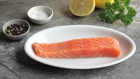 Video clip of food. A piece of fresh salmon on a white plate with Lemon, salt and pepper. Grey stone background. Real time.
