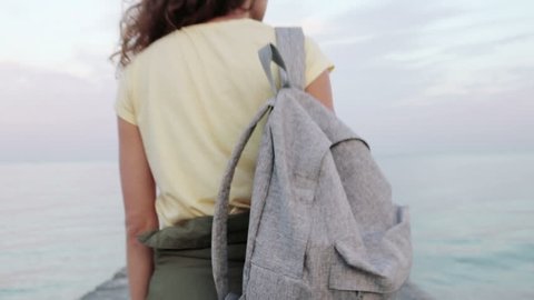 Young curly brunette in a yellow T-shirt with a backpack walking along the pier, stops at the edge and looks at the sea, view from the back, slow motion