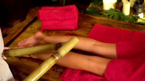 Bamboo feet massage massage with tapping. Young woman lying on wooden spa bed have bamboo massage therapy sticks. Bamboo massage in spa salon. Girl on candles background in massage spa salon.