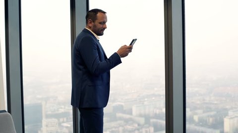 Young businessman standing with smartphone by window in the office
