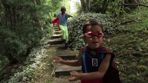 Two young boys dressed in homemade superhero costumes run through their backyard garden, jumping down stairs as they go. They through air punches and mimed energy blasts at the camera.