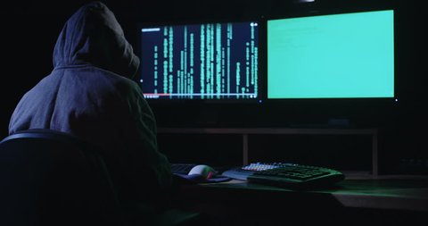 Computer hacker sitting in a dark room in front of computer screens hacking and writing lines of code