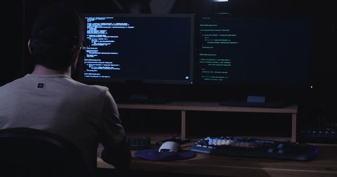 Computer hacker sitting in a dark room in front of computer screens hacking and writing lines of code