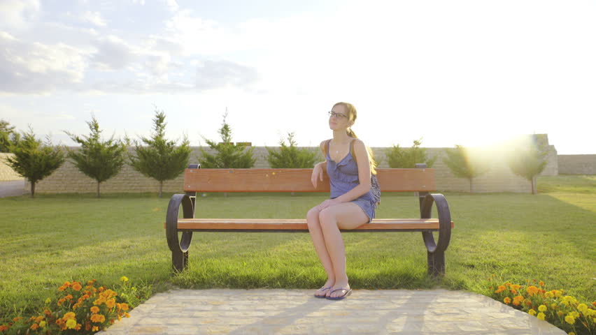 Woman enjoy green park while sitting on bench 4K. Camera moving around an attractive female person while sit and enjoy nature, green lawn around and sunshine in background.