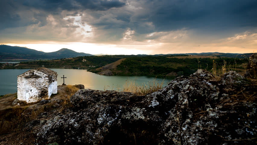 4K Tracking, panning and tilting timelapse of a church on hilltop over a big dam surrounded by mountains before big storm.