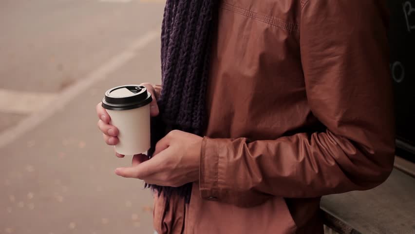 Young man with knitted scarf standing near the coffeeshop drinks coffee out of paper cup close up pan up