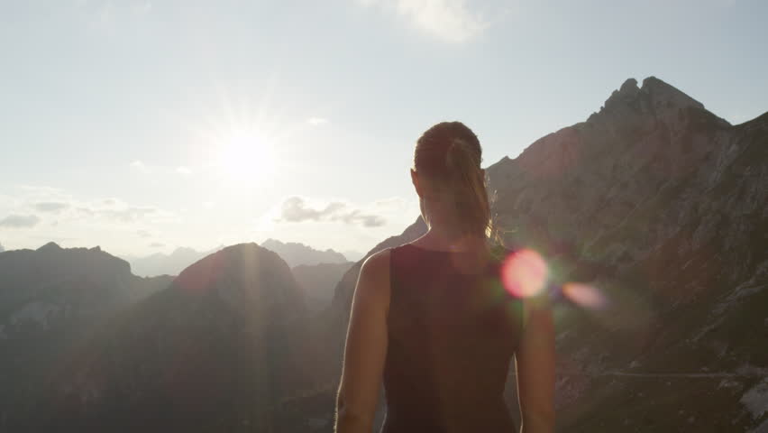 SLOW MOTION, CLOSE UP: Young female standing on the edge of the cliff and raising her hands up against high rocky mountains sunbathing in evening sun. Happy girl enjoying success and stunning view