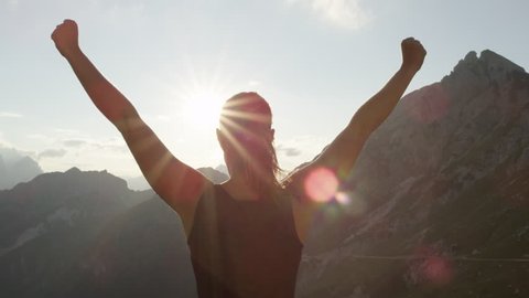 SLOW MOTION, CLOSE UP: Young female standing on the edge of the cliff and raising her hands up against high rocky mountains sunbathing in evening sun. Happy girl enjoying success and stunning view