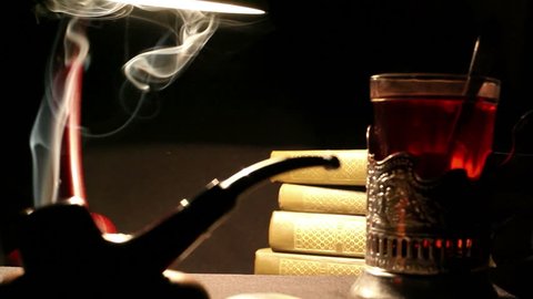 Night study interior: a person mixes sugar in a glass of tea in glass-holder, tobacco pipe smoke whirls, a pile of books lie beside under the lamplight
