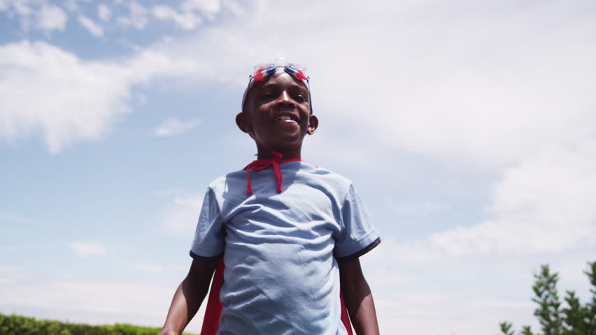 A young boy dressed in a homemade superhero costume raises his arms like a comic book hero. A golden light shines across him as he smiles. Bright, blue sky in the background. Low angle. Slow motion. Royalty-Free Stock Footage #20291719