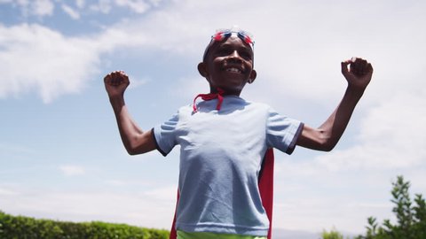 A young boy dressed in a homemade superhero costume raises his arms like a comic book hero. A golden light shines across him as he smiles. Bright, blue sky in the background. Low angle. Slow motion.