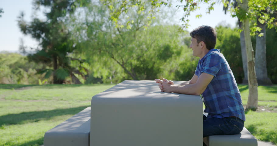 Young man waits at a concrete table and bench at a park in Los Angeles.  Medium long shot, recorded hand-held in real time from side angle.